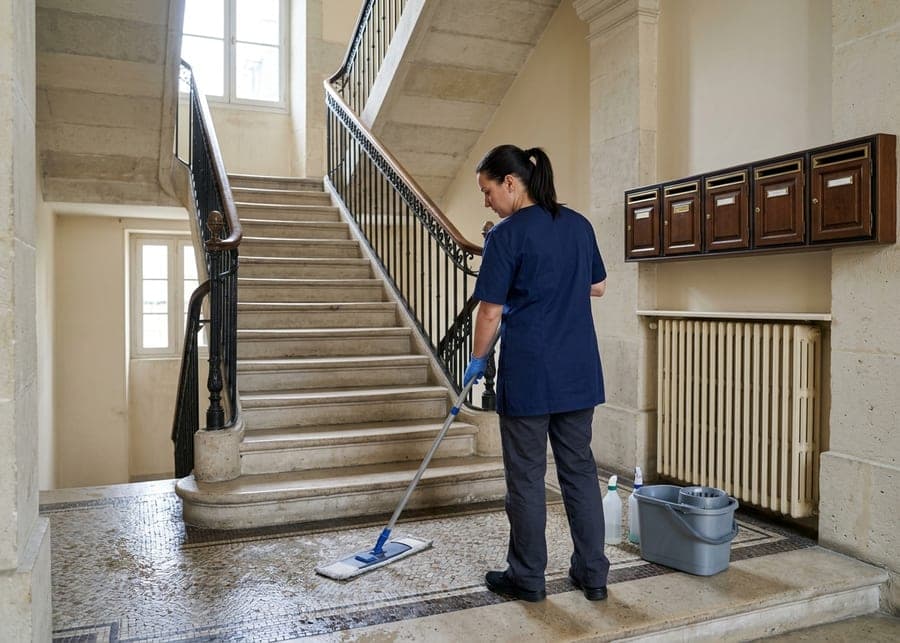 Femme qui lave au balai le palier carrelé d’un hall d’immeuble en pierre, escalier et boîtes aux lettres